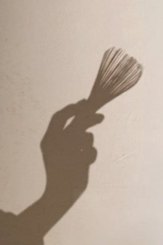 The shadow of a hand holding a bamboo matcha whisk against a warm wall